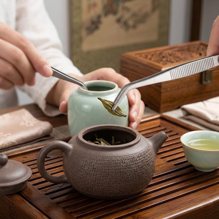 Stainless steel tea tongs lifting tea leaves above a green kettle