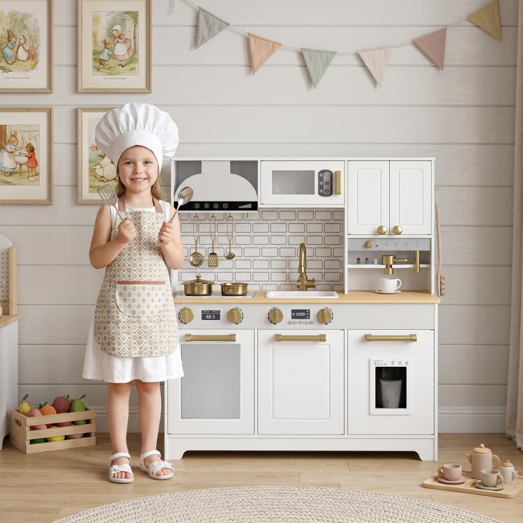 Child in chef hat and apron standing by white kids play kitchen with gold faucet