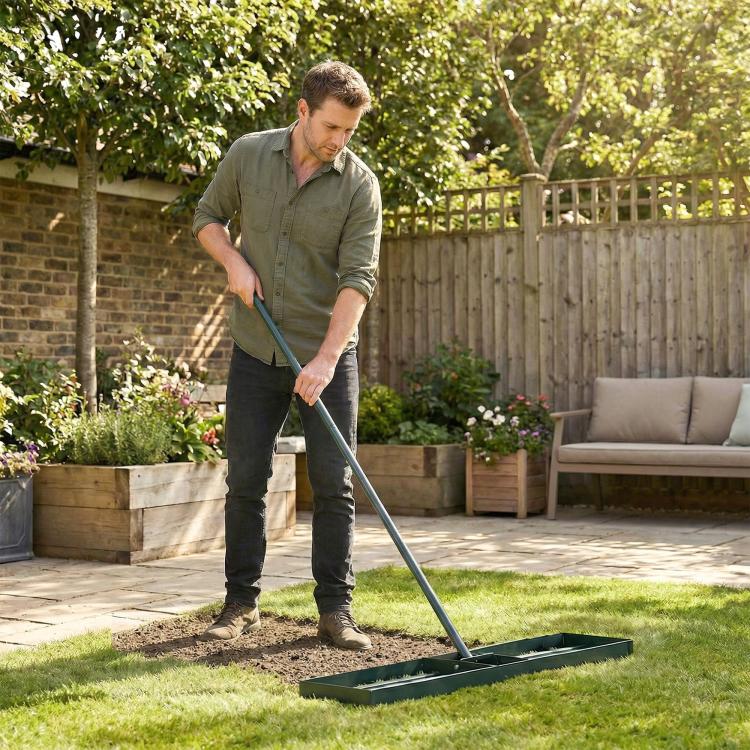Green leveling rake spreading soil across lawn, held by a gardener