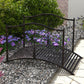 Outdoor dark bronze arched garden bridge with scroll side rails over gravel pathway.