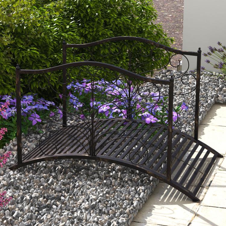 Outdoor dark bronze arched garden bridge with scroll side rails over gravel pathway.