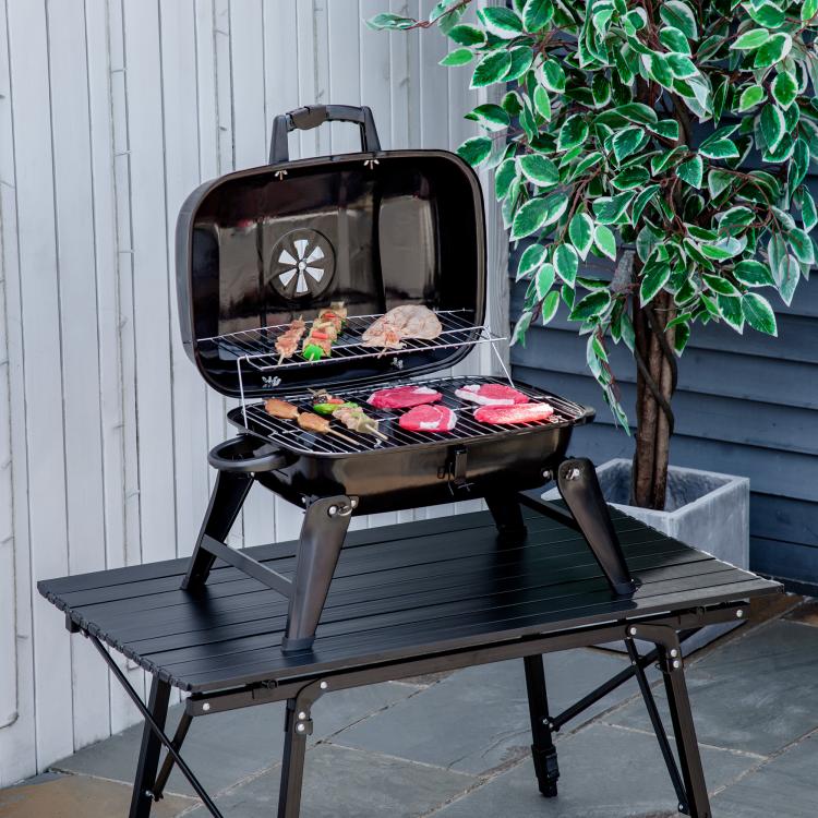Black folding portable charcoal grill on an outdoor table, lid open with food on grates