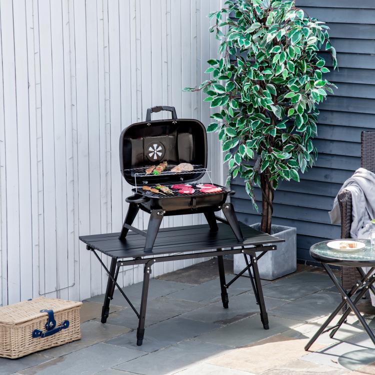 Black portable charcoal grill with lid open, food cooking on top grate outdoors