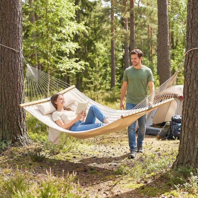Cream two-person outdoor hammock with pillow and pad, stretched between wooden spreaders