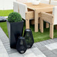 Tall black planter with dense green plant outdoors beside woven patio furniture