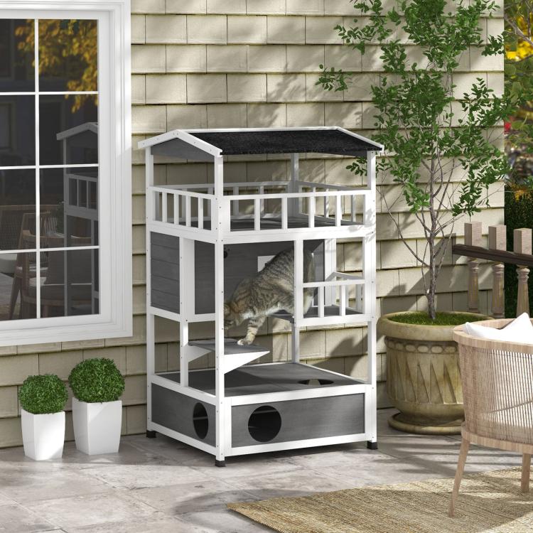 Light gray wooden cat house with dark roof and side entrance opening, outdoors