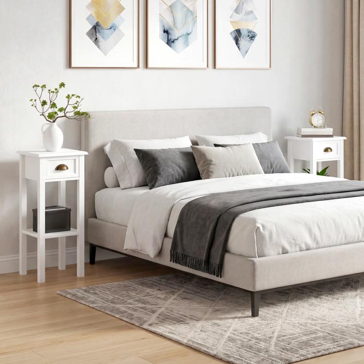 White 2-tier nightstand-style side table with drawer and open shelf beside a bed.