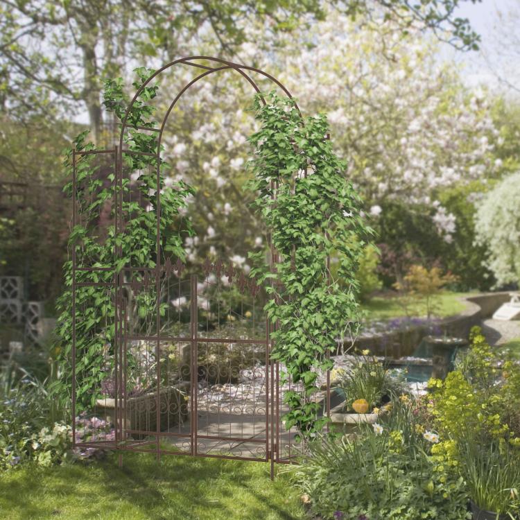 Brown metal garden arch arbor gate set among green shrubs and flowers in a yard