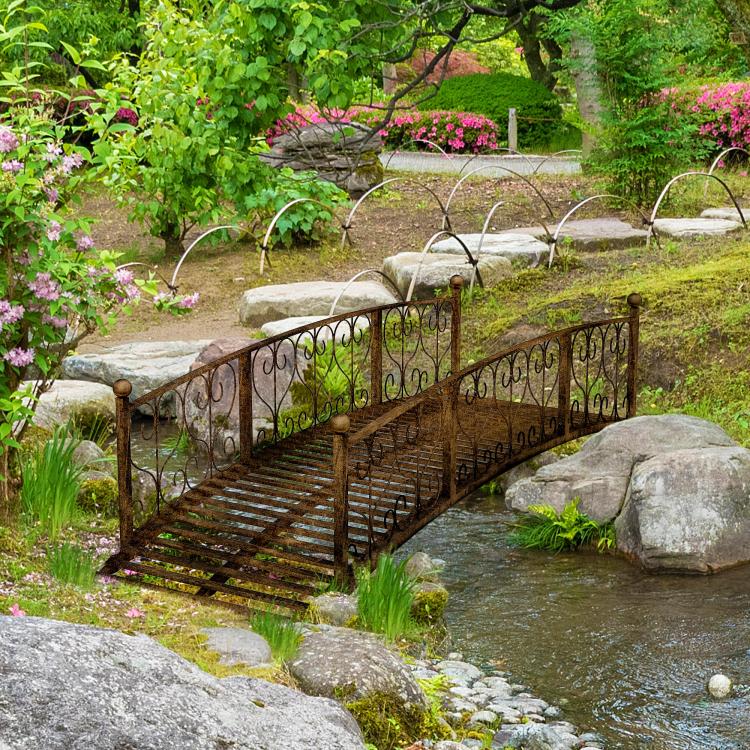 Bronze metal garden footbridge spanning a small stream with scroll side railings