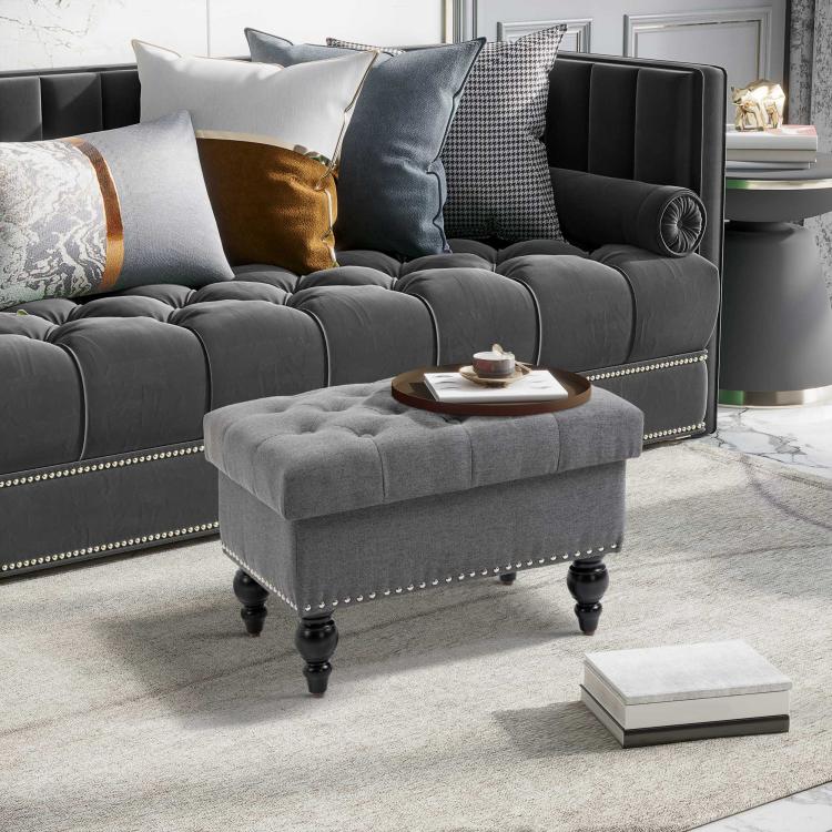 Gray storage ottoman with tufted seat, nailhead trim edge, and turned legs