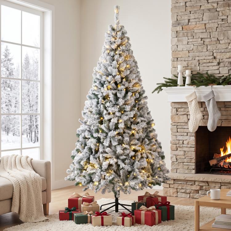 Snow-flocked prelit Christmas tree with full silhouette and warm lights, standing on base.