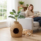 Rattan-style cat litter box with faux plant top and round front doorway next to orange cat