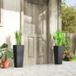 Two black tall woven planters with potted plants in a courtyard beside a wall