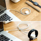 White magnetic charging puck and cable on wooden desk beside laptop and watch