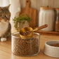 Stainless steel pet food measuring scoop with wooden handle over a glass kibble jar