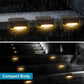 Black solar stair lights in rainfall, warm LEDs glowing on steps and near the wall.