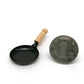Miniature black frying pan with wooden handle, shown with coin size reference