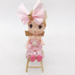 Doll in pink outfit seated on miniature wooden dollhouse chair.