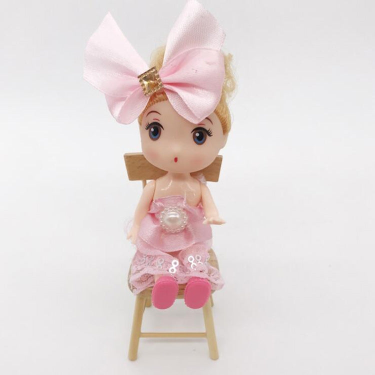 Doll in pink outfit seated on miniature wooden dollhouse chair.