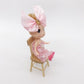 Doll seated on miniature wooden dollhouse chair with slatted back.