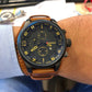 Men's quartz watch on wrist with brown leather band and black dial, yellow markers and subdials.