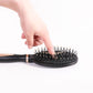 Black hair massage brush being held, showing dense silver pins on the oval pad.