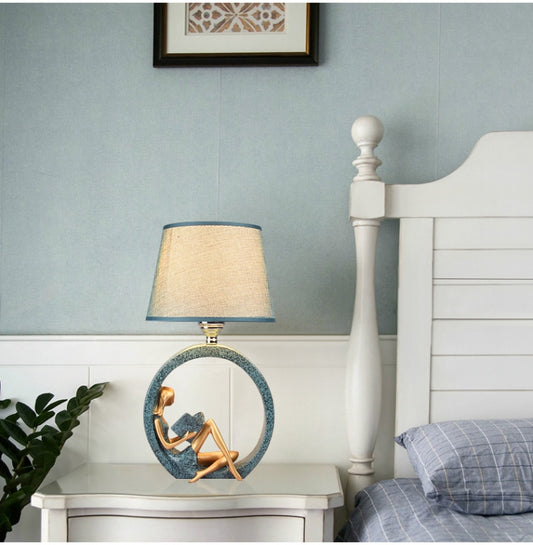 Bedside table lamp with blue fabric shade and circular figurine base on a nightstand.