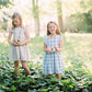 Wooden camera toy with round lens, held by child; light wood and blue plaid dress