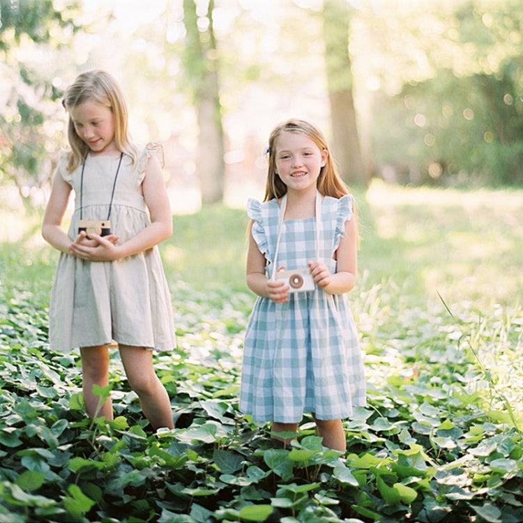 Wooden camera toy with round lens, held by child; light wood and blue plaid dress