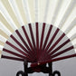 White blank silk folding fan with dark maroon radial stripes and dark red inner circle on a stand.