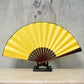 Yellow blank silk folding fan with maroon inner ribs, Chinese-style, on black stand