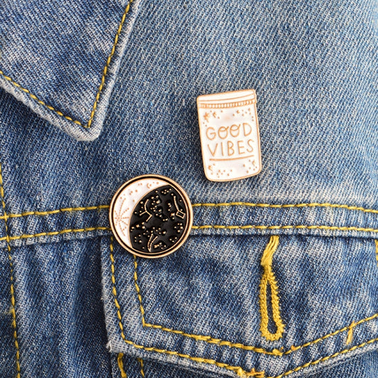 Enamel brooch pins on denim: round moon constellation and “Good Vibes” bottle pin.