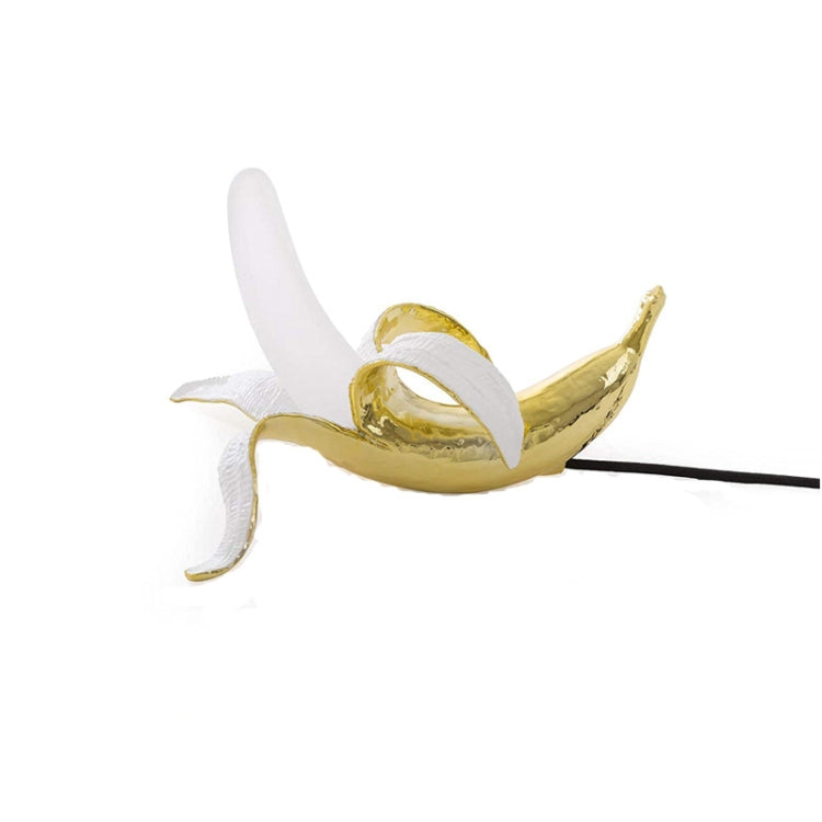 Banana-shaped table lamp with gold banana body and white bulb, stretched pose