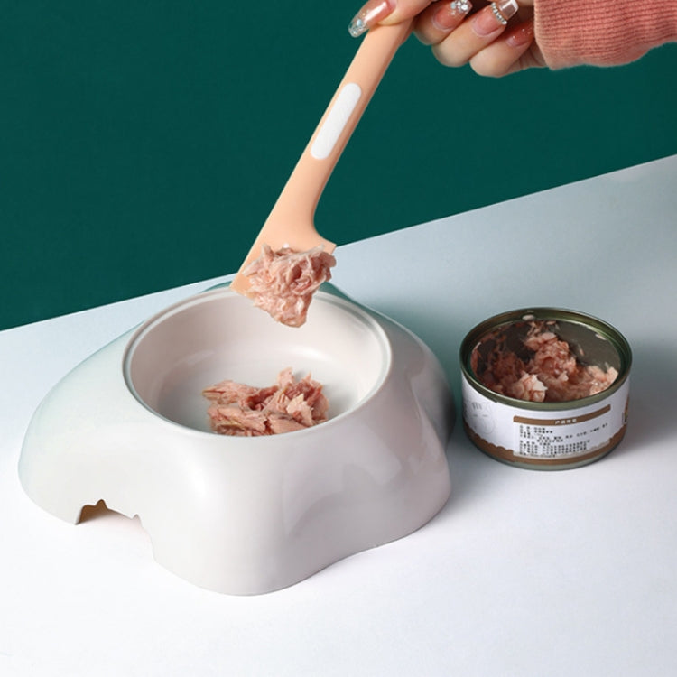 Pink cat-shaped spoon lifting wet pet food; metal bowl with small pet can.