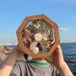 Wooden kaleidoscope toy with octagon viewing window held up by two hands by sea