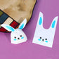 Rabbit-ear gift bag, white with blue ears and cartoon cat/bunny face, tied closed.