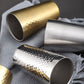 Hammer-finish stainless steel mugs in gold and silver, angled on cloth