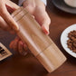 Light wooden hand coffee grinder with crank handle, held by two hands