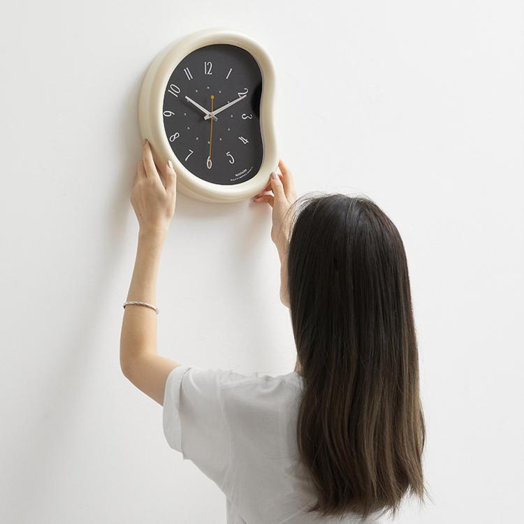 Beige-framed wall clock being held for mounting, dark face with number markers