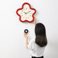 Flower-shaped wall clock with red frame, cream face, black hands; hand adjusting it.
