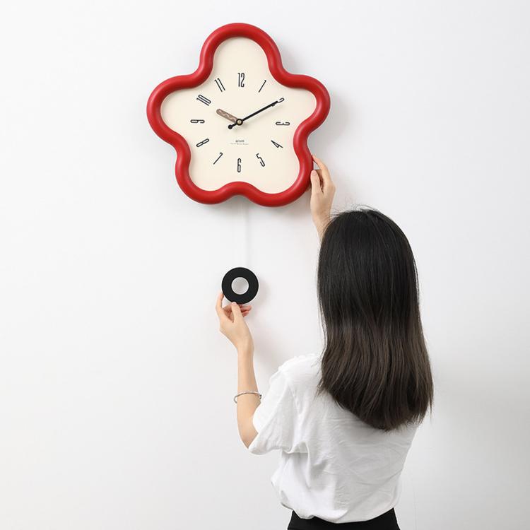 Flower-shaped wall clock with red frame, cream face, black hands; hand adjusting it.