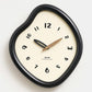 Irregular wall clock with glossy dark frame and cream dial, with gold-toned hands.
