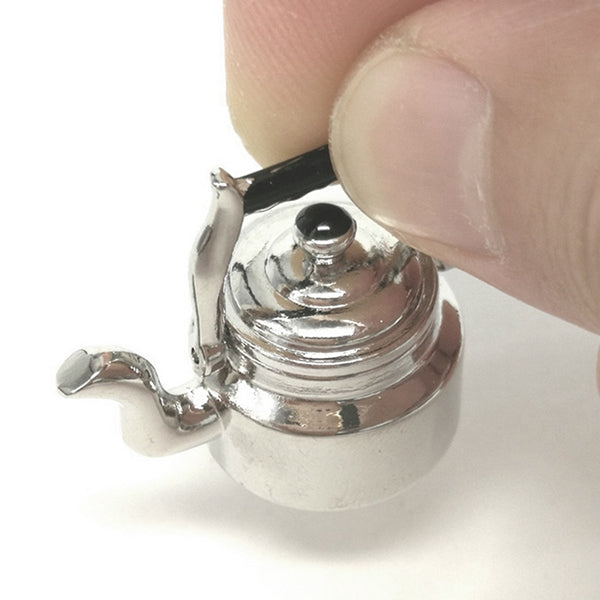 Miniature metal tea kettle with black knob lid, silver spout, held by a finger.