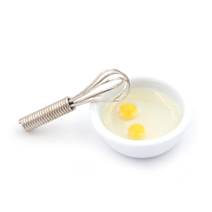 Miniature manual egg beater with metal whisk and small bowl of two eggs