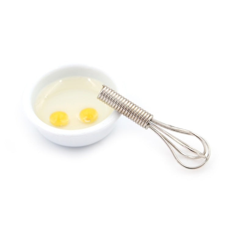 Miniature manual egg beater with metal whisk next to small bowl with two eggs