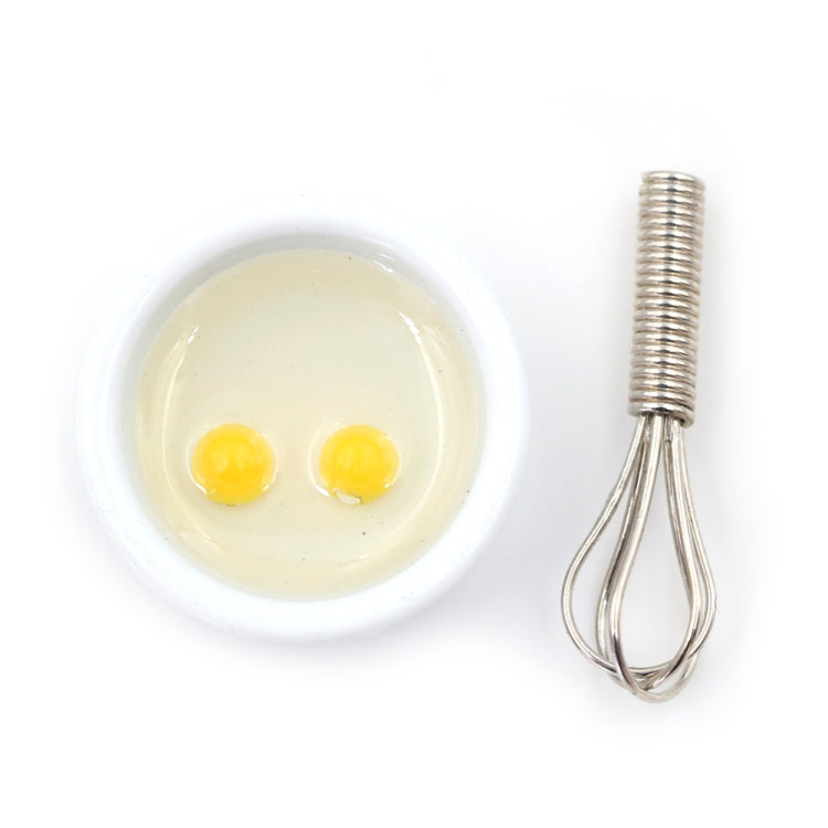 Miniature manual egg beater with metal whisk and small white bowl containing two eggs